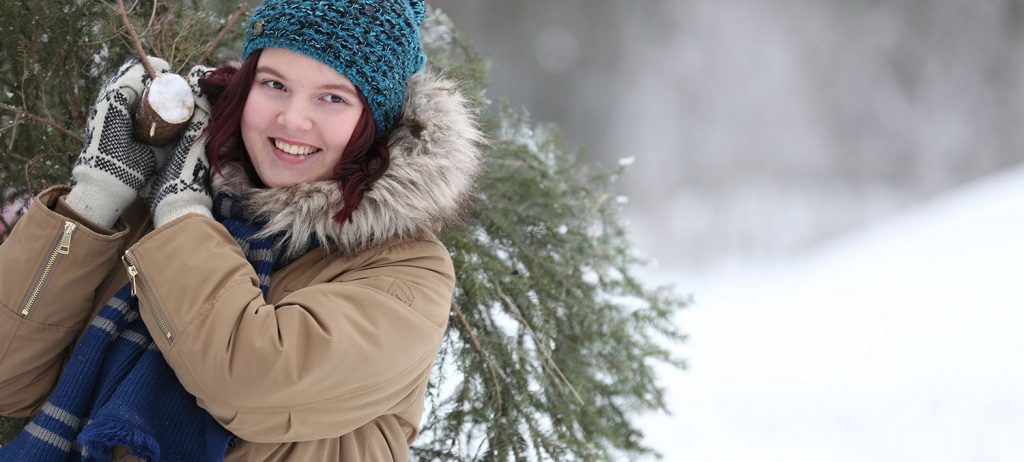 Nainen kantaa hymyillen joulukuusta olallaan talvisessa maisemassa