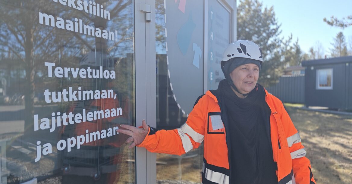 Mari Juntunen nojaa lasiseinäiseen konttiin, jossa asiakkaat voivat testata uutta tapaa lajitella poistotekstiiliä.